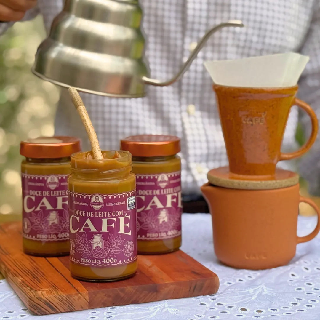 Potes de Doce de Leite com Café Dona Lázara 400g ao lado de um café sendo coado em coador de cerâmica, com xícara e bule