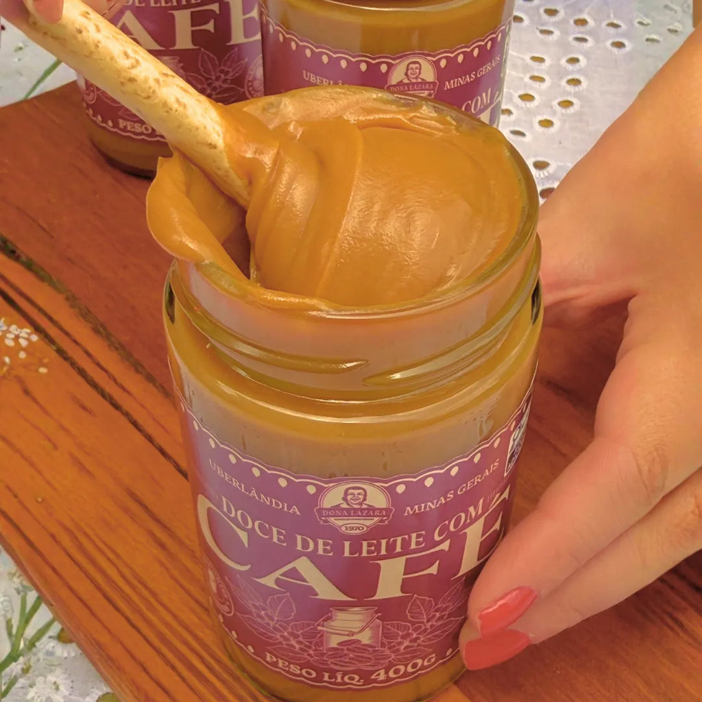 Mão segurando um pote aberto de Doce de Leite com Café Dona Lázara 400g, com um palito puxando o doce cremoso