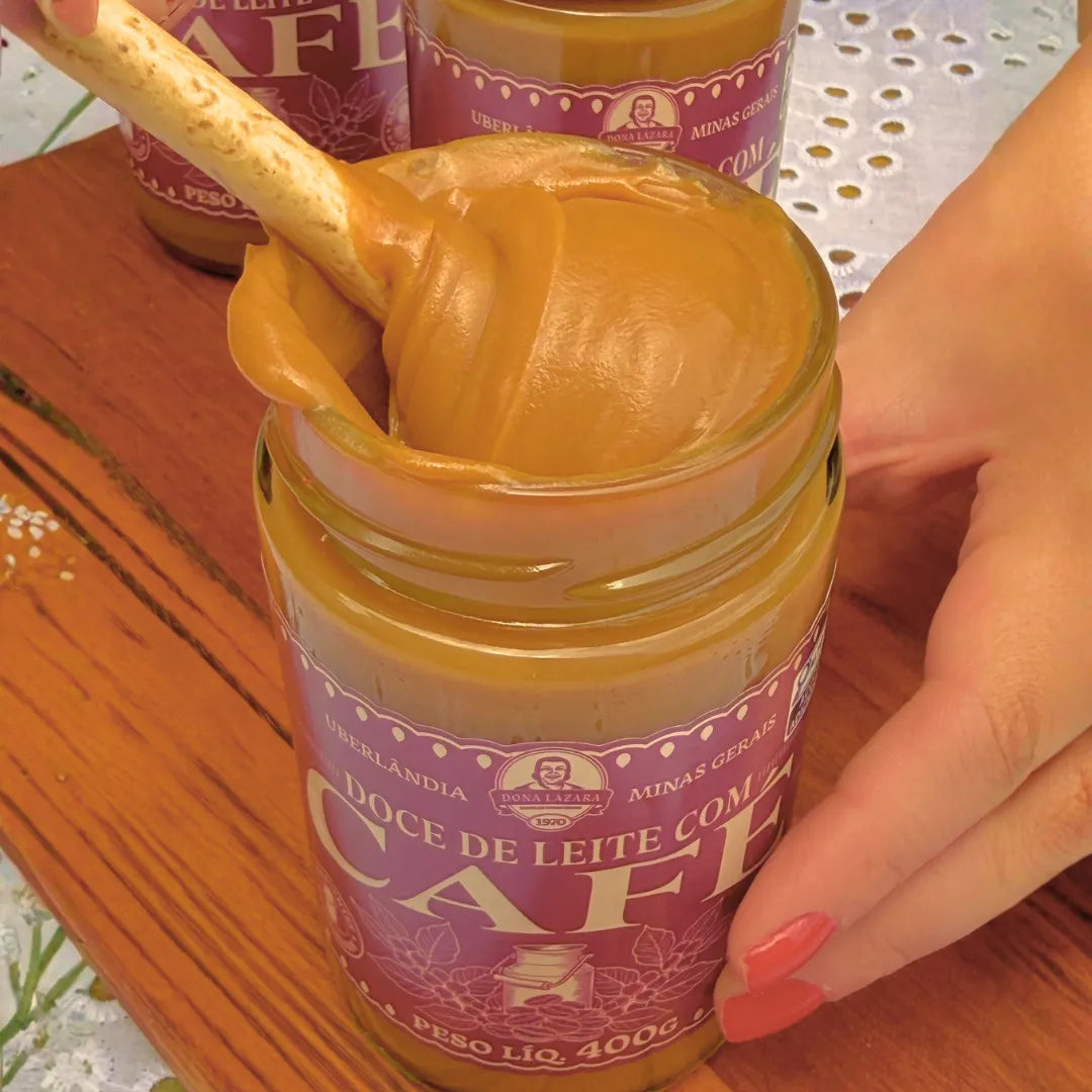 Mão segurando um pote aberto de Doce de Leite com Café Dona Lázara 400g, com um palito puxando o doce cremoso