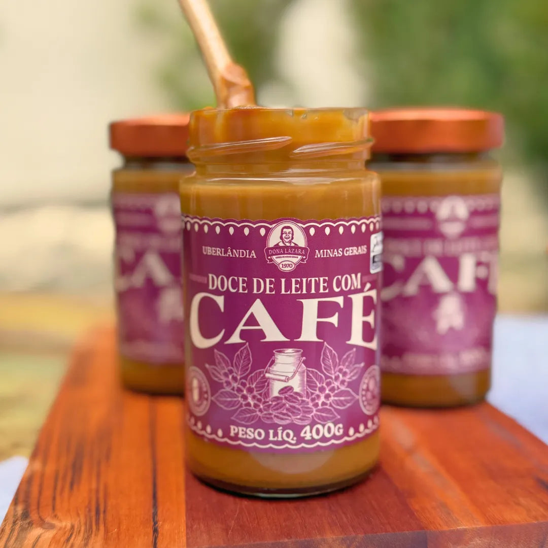 Três potes de Doce de Leite com Café Dona Lázara 400g em tábua de madeira, o da frente aberto com colher de pau