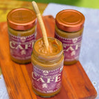 Três potes de Doce de Leite com Café Dona Lázara 400g de tampa cobre em tábua de madeira, o da frente aberto com a colher espetada no doce