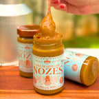 Pote aberto de Doce de Leite com Nozes Dona Lázara 400g, com uma colherada do doce cremoso sendo erguida, ao ar livre
