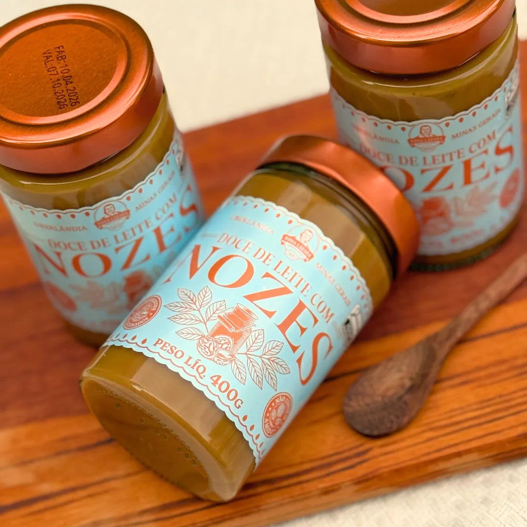 Três potes de Doce de Leite com Nozes Dona Lázara 400g sobre tábua de madeira, um deitado mostrando a tampa cobre, com colher de pau ao lado