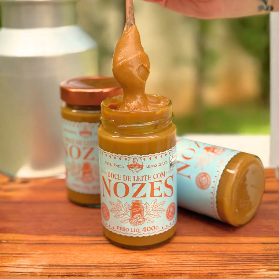 Pote aberto de Doce de Leite com Nozes Dona Lázara 400g com colher mergulhada no doce cremoso, leiteira e potes ao fundo no jardim