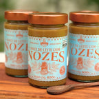 Três potes de Doce de Leite com Nozes Dona Lázara 400g em pé sobre tábua de madeira, com a lupa "Alto em açúcar adicionado" no rótulo e colher de pau ao lado