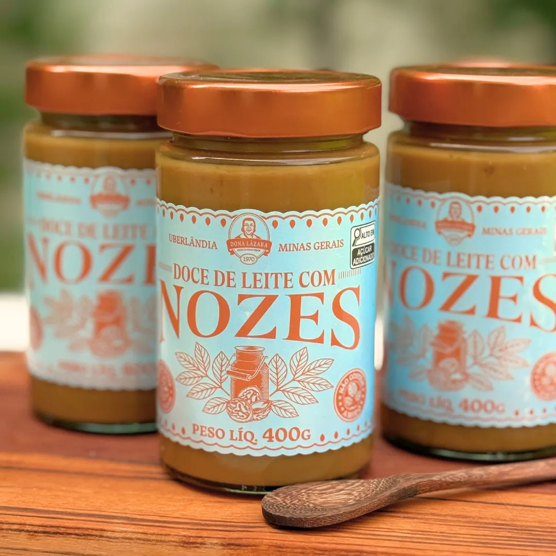 Três potes de Doce de Leite com Nozes Dona Lázara 400g em pé sobre tábua de madeira, com a lupa "Alto em açúcar adicionado" no rótulo e colher de pau ao lado