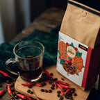 Pacote do café Especiarias ao lado de uma xicara de vidro com café e pimentas e grãos ao redor.