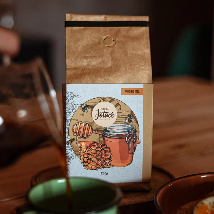 Pacote de Café Favo de Mel da Jotacê com jarra e xícara à frente em uma mesa de madeira
