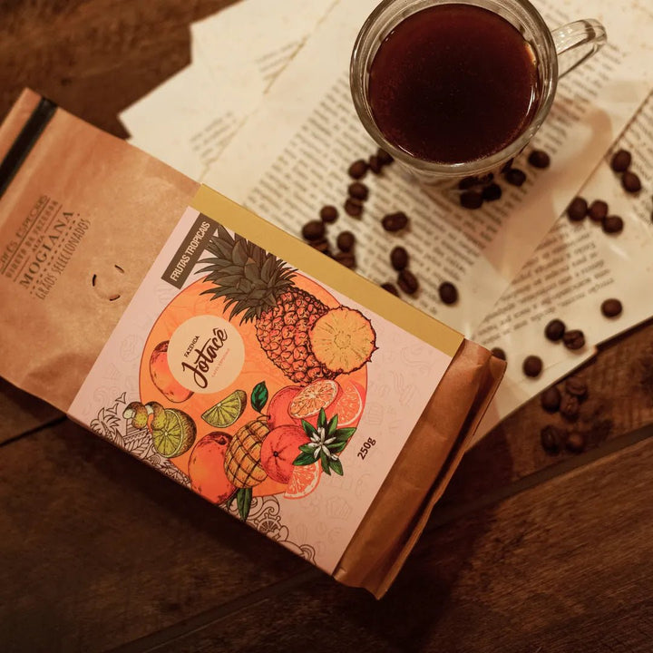 Pacote do Café Frutas Tropicais sobre folhas rasgadas de um livro ao lado de uma xícara de café e grãos espalhados