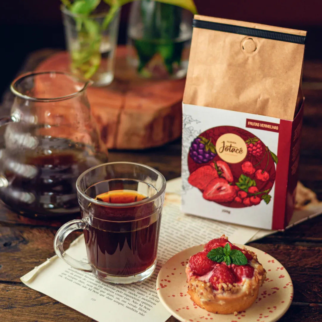Xícara de café Frutas Vermelhas servida ao lado de uma torta de morango e a embalagem do café sobre mesa de madeira
