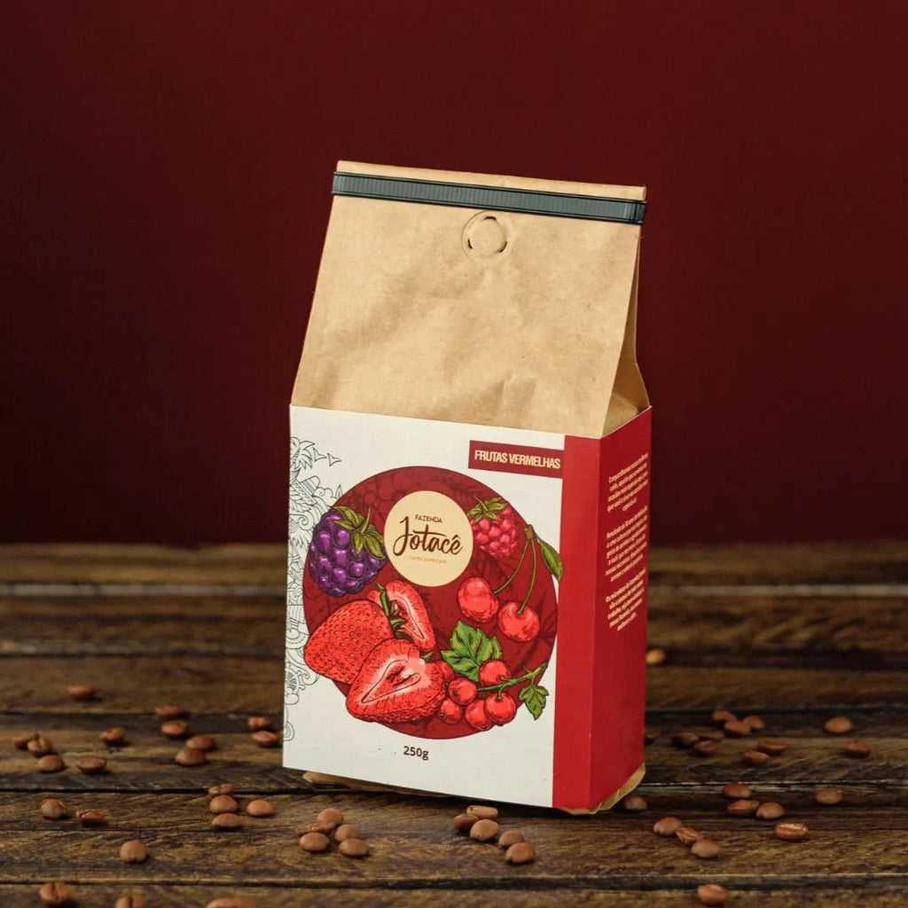 Pacote de café Frutas Vermelhas da Fazenda Jotacê sobre mesa rústica com grãos de café torrado espalhados.