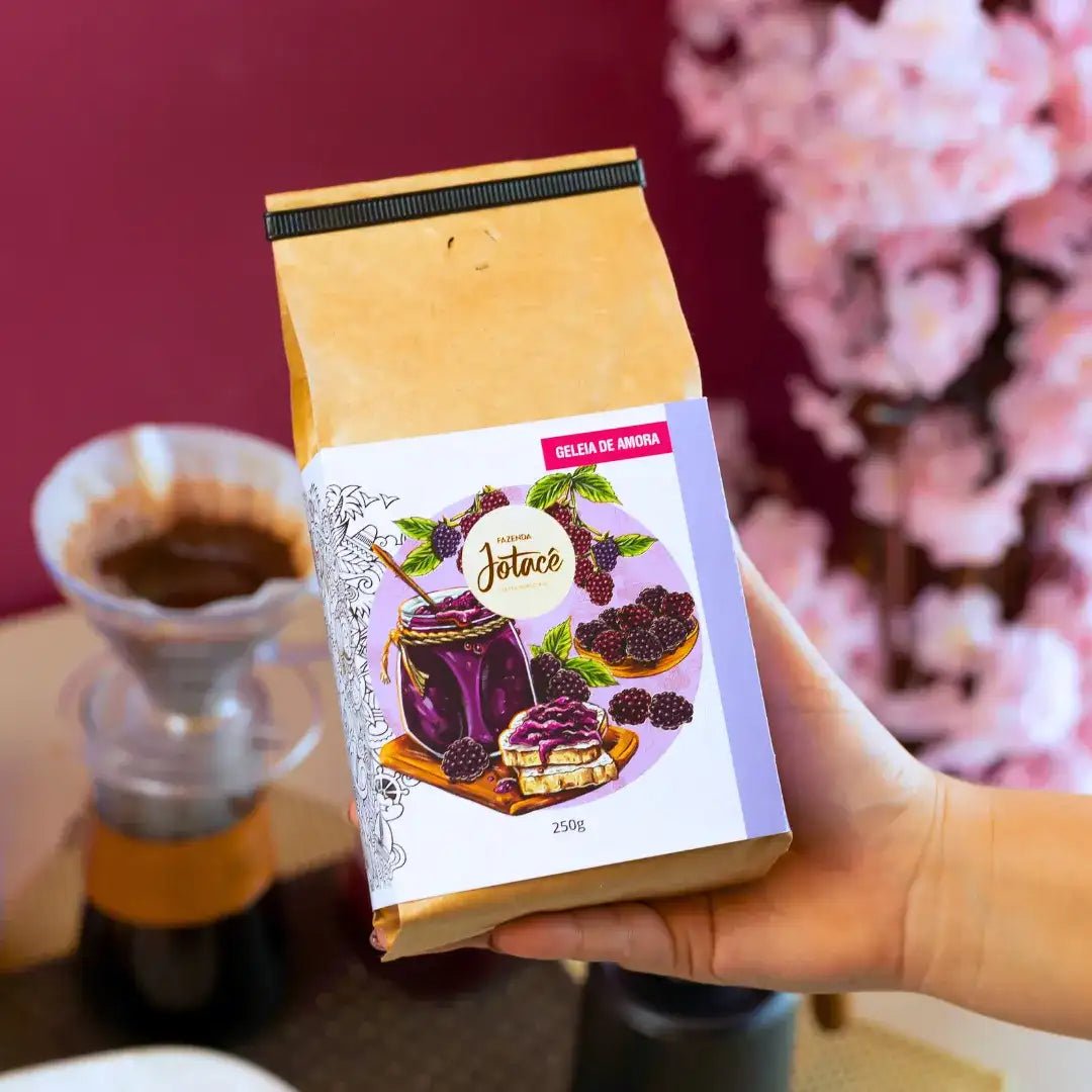 Mão segurando um pacote de Café Especial Geleia de Amora da Fazenda Jotacê, com fundo em tons quentes e um coador Hario V60 ao fundo, ressaltando a experiência afetiva e nostálgica desse microlote.