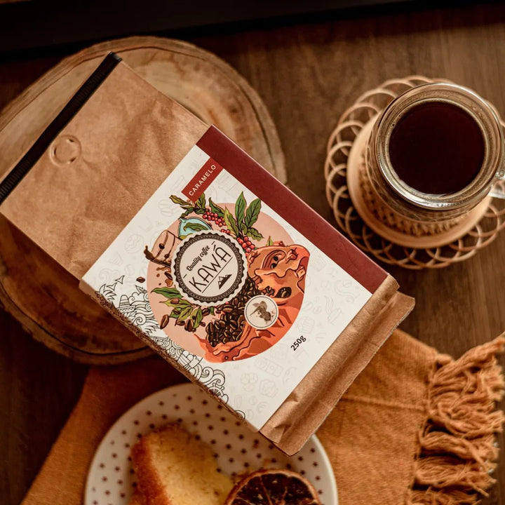 Vista superior do Pacote de 250g do café Kawá Caramelo da Fazenda Jotacê, ao lado de um bolo e xícara sobre mesa de madeira