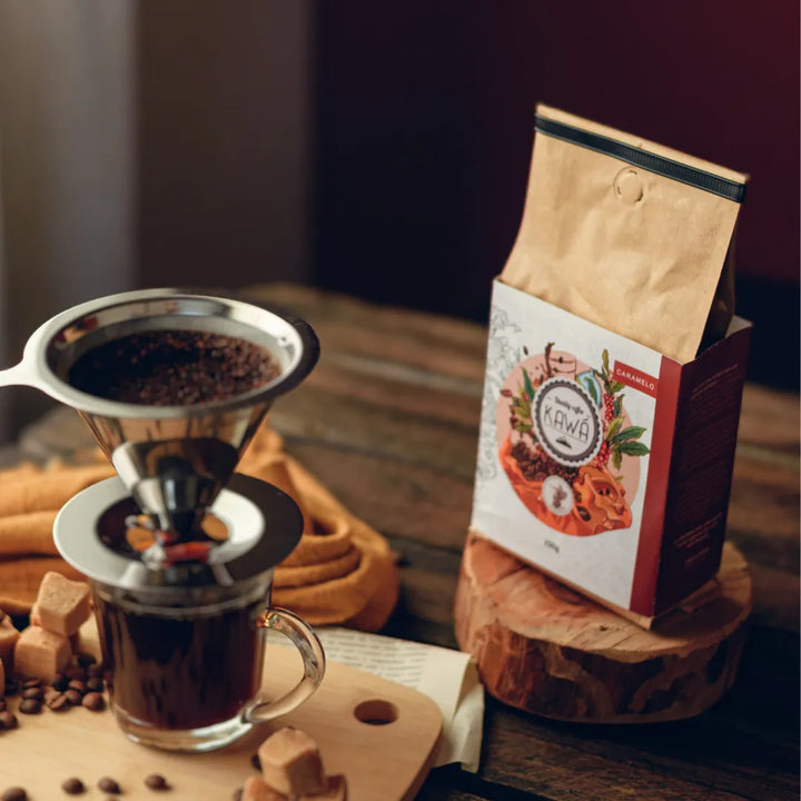Pacote do Café Kawá Caramelo da Fazenda Jotacê ao lado de um filtro inox extraindo café em uma xicara sobre mesa de madeira