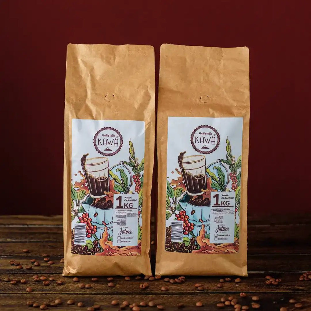 2 pacotes de 1 Kg em pé do produto Kawá Caramelo dispostos lateralmente sobre mesa de madeira com grãos soltos