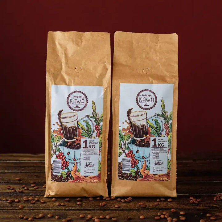 2 pacotes de 1 Kg em pé do produto Kawá Caramelo dispostos lateralmente sobre mesa de madeira com grãos soltos