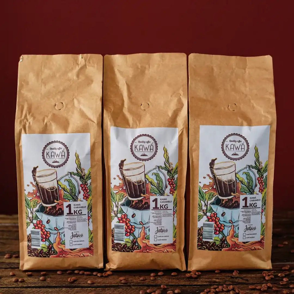 3 pacotes de 1 Kg em pé do produto Kawá Caramelo dispostos lado a lado sobre mesa de madeira com grãos soltos