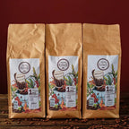 3 pacotes de 1 Kg em pé do produto Kawá Caramelo dispostos lado a lado sobre mesa de madeira com grãos soltos