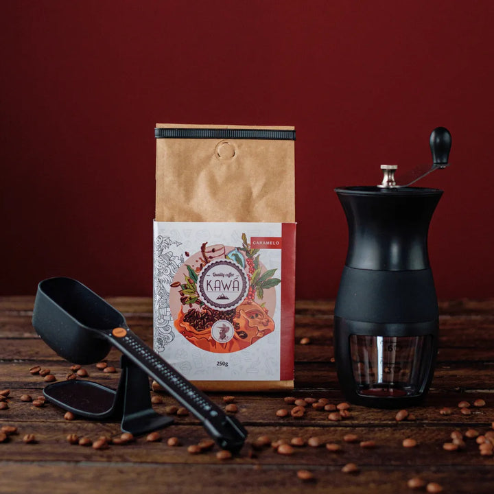 Kit de café com moedor manual, pacote de café Kawá e balança de dose, dispostos sobre superfície de madeira.
