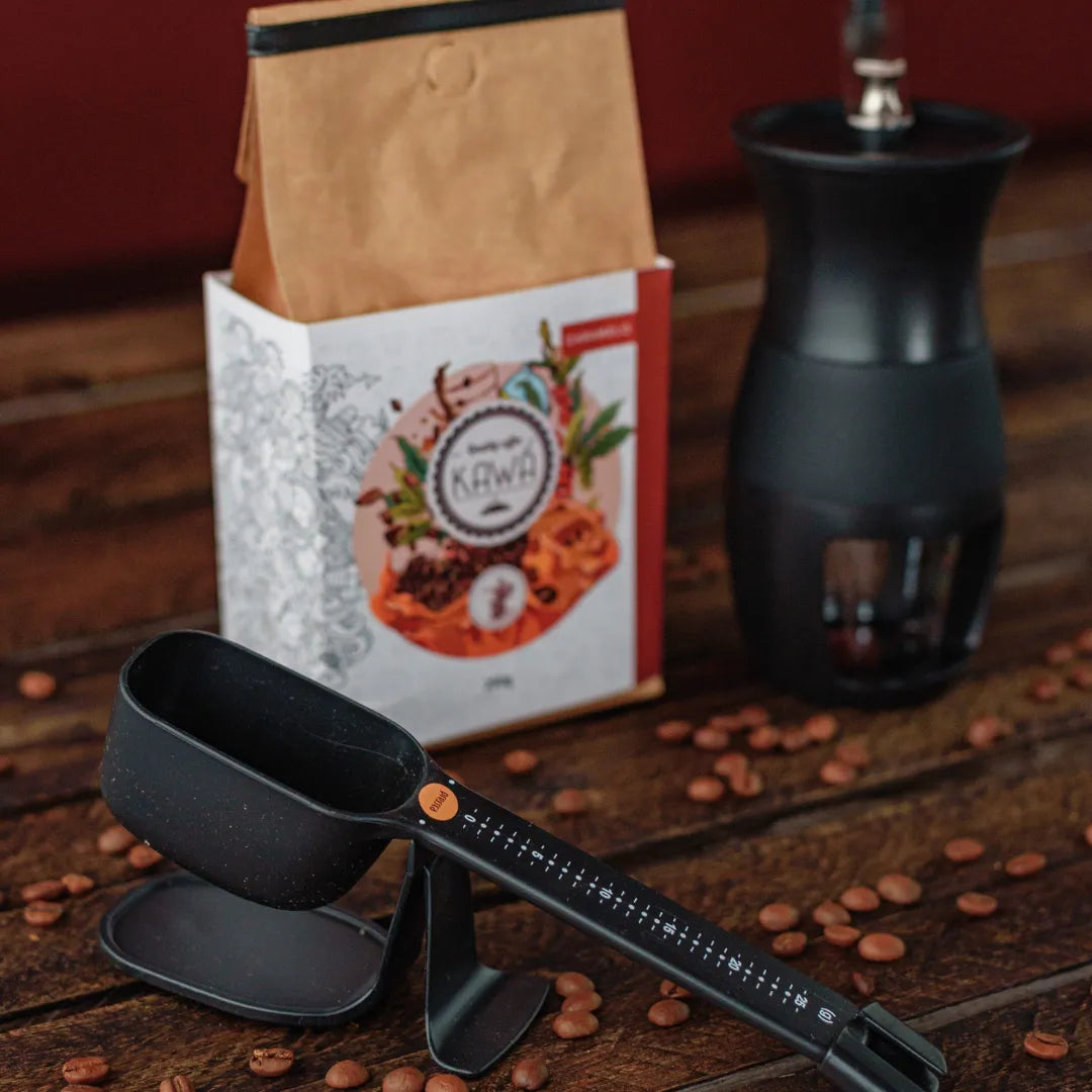 Acessórios para café com moedor, balança, e pacote de café Kawá sobre uma mesa de madeira.