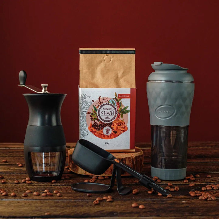 Equipamento de café com moedor manual, pacote de café Kawá, balança e cafeteira portátil Pressca, em uma mesa de madeira.