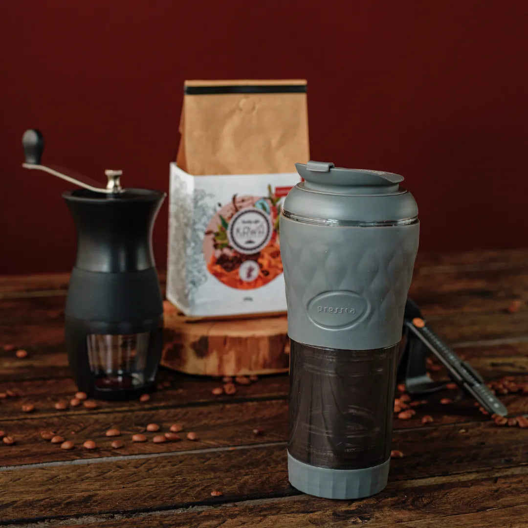 Equipamento de café com moedor manual, pacote de café Kawá, e cafeteira portátil Pressca, em uma mesa de madeira.