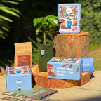 Kit Coleção Raízes da Fazenda Jotacê sobre tronco de madeira ao ar livre: caixa azul, pillow box com sementes de café arábica, terra TERRAL, vaso King Pot com muda de café e revista guia.