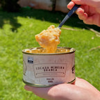 Mão segurando lata aberta de Cocada Mineira Branca Dona Lázara, com colher erguendo o doce cremoso sob o sol, mostrando sua textura artesanal e brilhante.