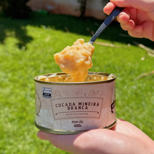 Mão segurando lata aberta de Cocada Mineira Branca Dona Lázara, com colher erguendo o doce cremoso sob o sol, mostrando sua textura artesanal e brilhante.