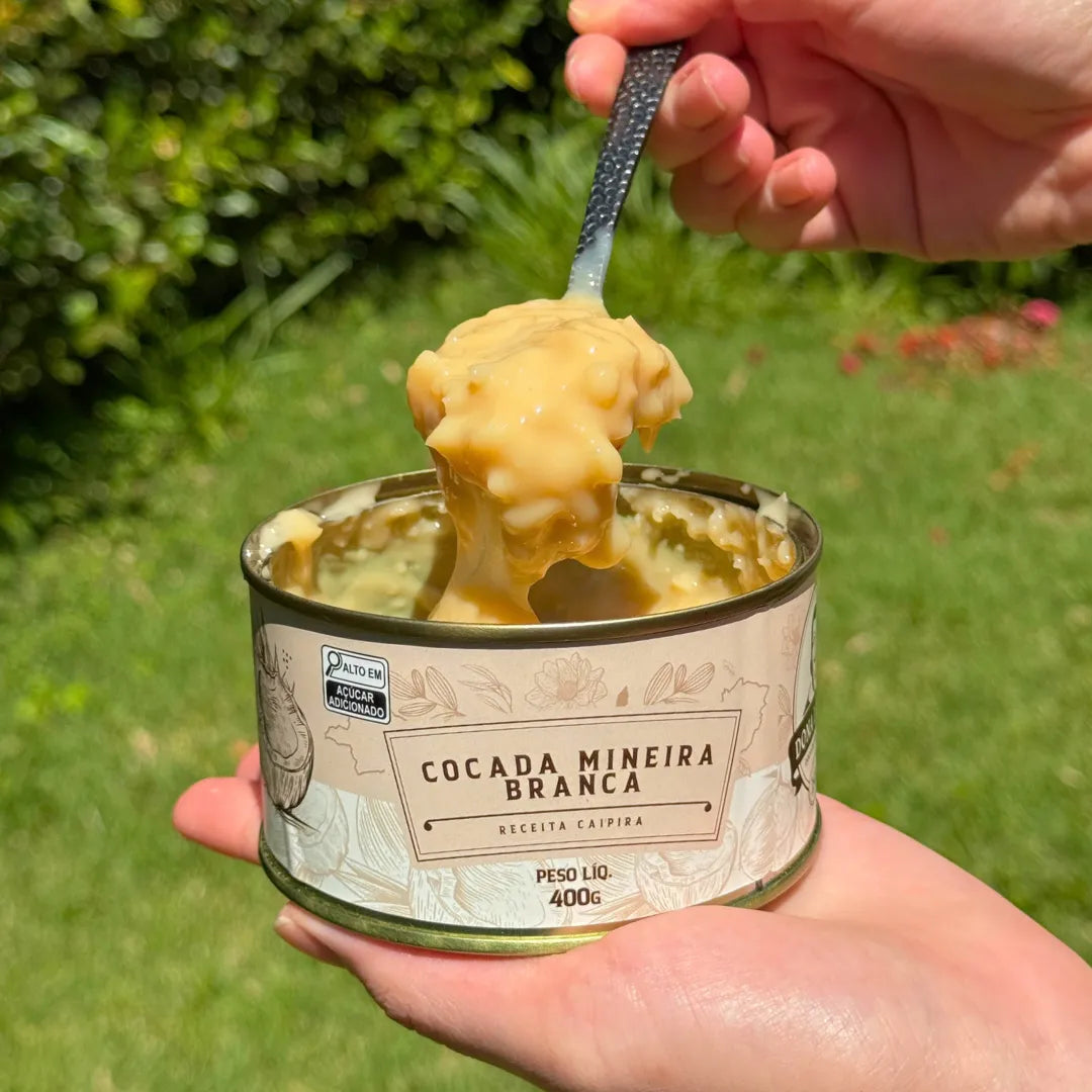 Colher mergulhando na Cocada Mineira Branca Dona Lázara, revelando a cremosidade do doce e o coco fresco, com latas e cocos compondo o cenário.