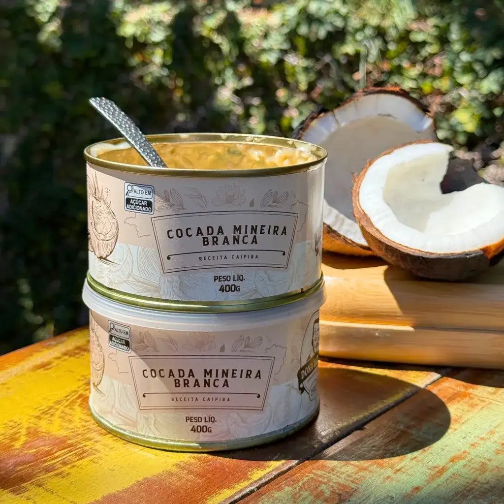 Duas latas de Cocada Mineira Branca Dona Lázara empilhadas, com coco partido ao fundo, sobre mesa rústica de madeira, em ambiente ensolarado.