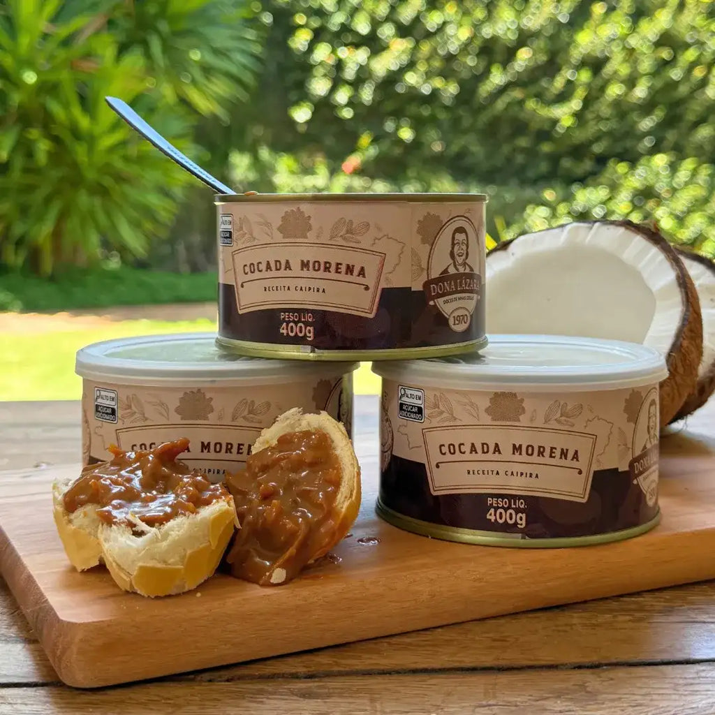 Lata de Cocada Morena 400g aberta sobre uma tábua, com pedaços de pão recheados com a cocada, e outras latas ao fundo ao lado de um coco partido.