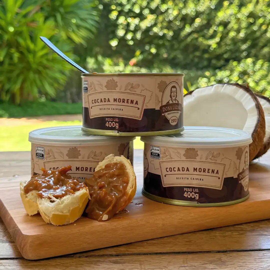 Lata de Cocada Morena 400g aberta sobre uma tábua, com pedaços de pão recheados com a cocada, e outras latas ao fundo ao lado de um coco partido.