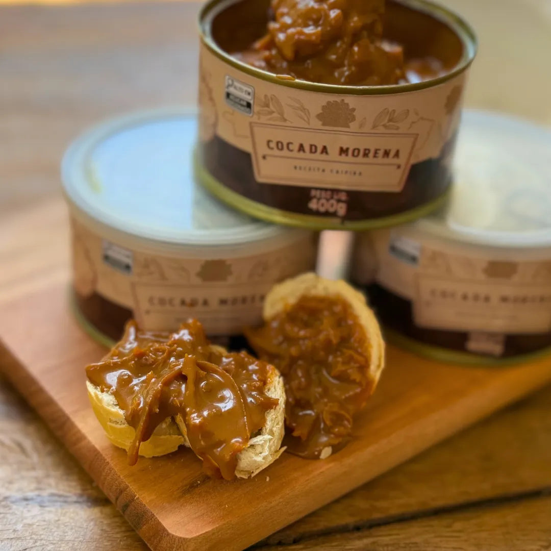 Close da Cocada Morena 400g aberta, com pedaços de coco fresco ao fundo e a cocada espalhada sobre fatias de pão em uma tábua de madeira.