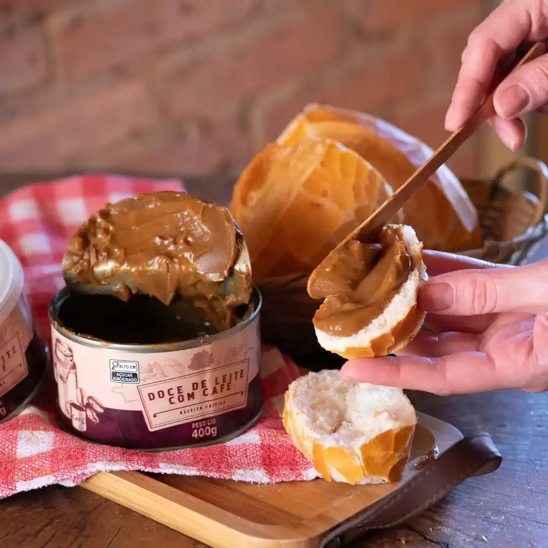 Pessoa passando doce de leite com café em uma fatia de pão francês. A lata aberta está ao fundo, junto com uma cesta cheia de pães e um pano xadrez vermelho e branco.