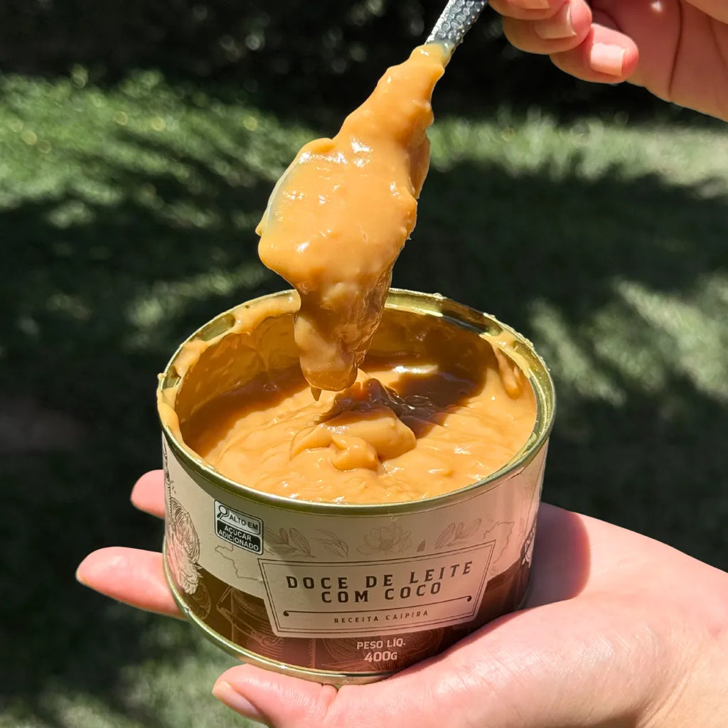 Lata aberta de Doce de Leite com Coco 400g, mostrando a textura cremosa do doce com colher apoiada na borda.