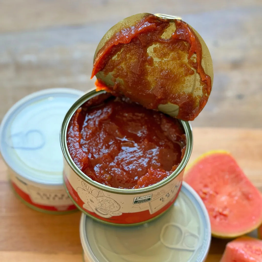 Três latas de goiabada cremosa e fatias de goiaba fresca com queijo minas sobre tábua de madeira.