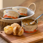 Lata de Doce de Leite Receita Caipira 400g aberta ao lado de pães de queijo recheados com o doce, em tábua de madeira.