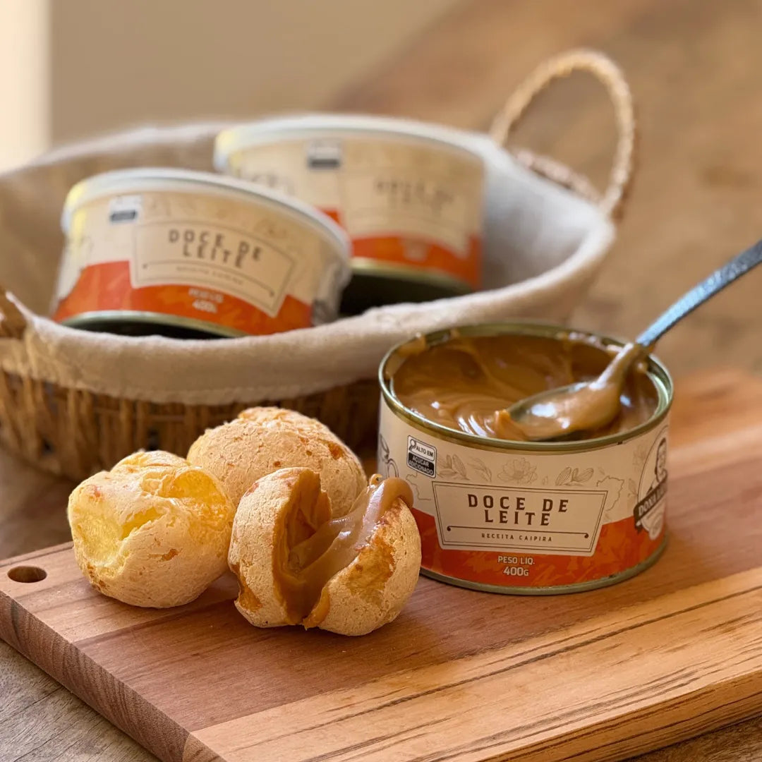 Lata de Doce de Leite Receita Caipira 400g aberta ao lado de pães de queijo recheados com o doce, em tábua de madeira.
