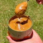 Doce de Leite Receita Caipira 400g sendo puxado com uma colher, mostrando sua cremosidade, com a lata apoiada na mão.