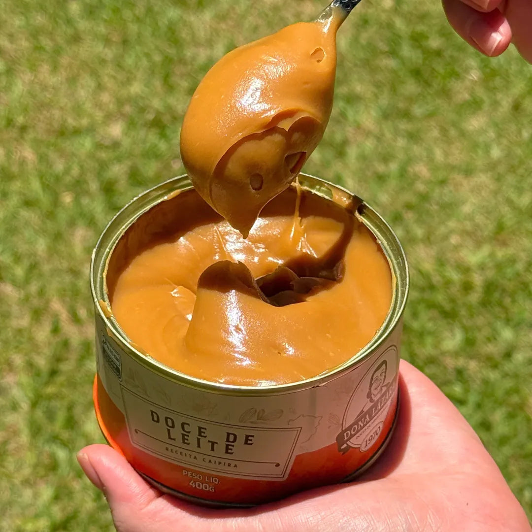 Doce de Leite Receita Caipira 400g sendo puxado com uma colher, mostrando sua cremosidade, com a lata apoiada na mão.