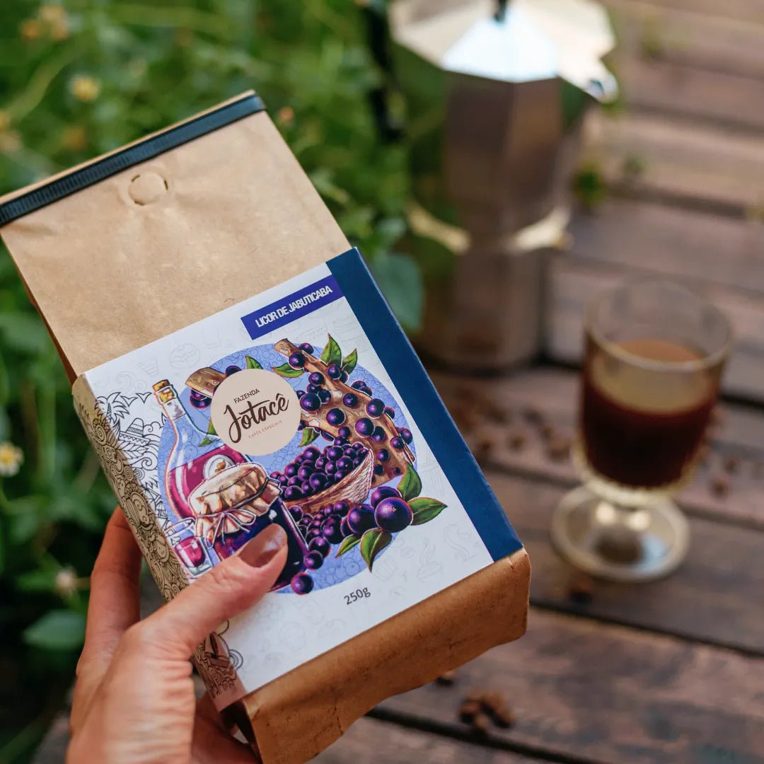 "Mão segurando embalagem de Café Licor de Jabuticaba com xícara e cafeteira Italiana ao fundo.