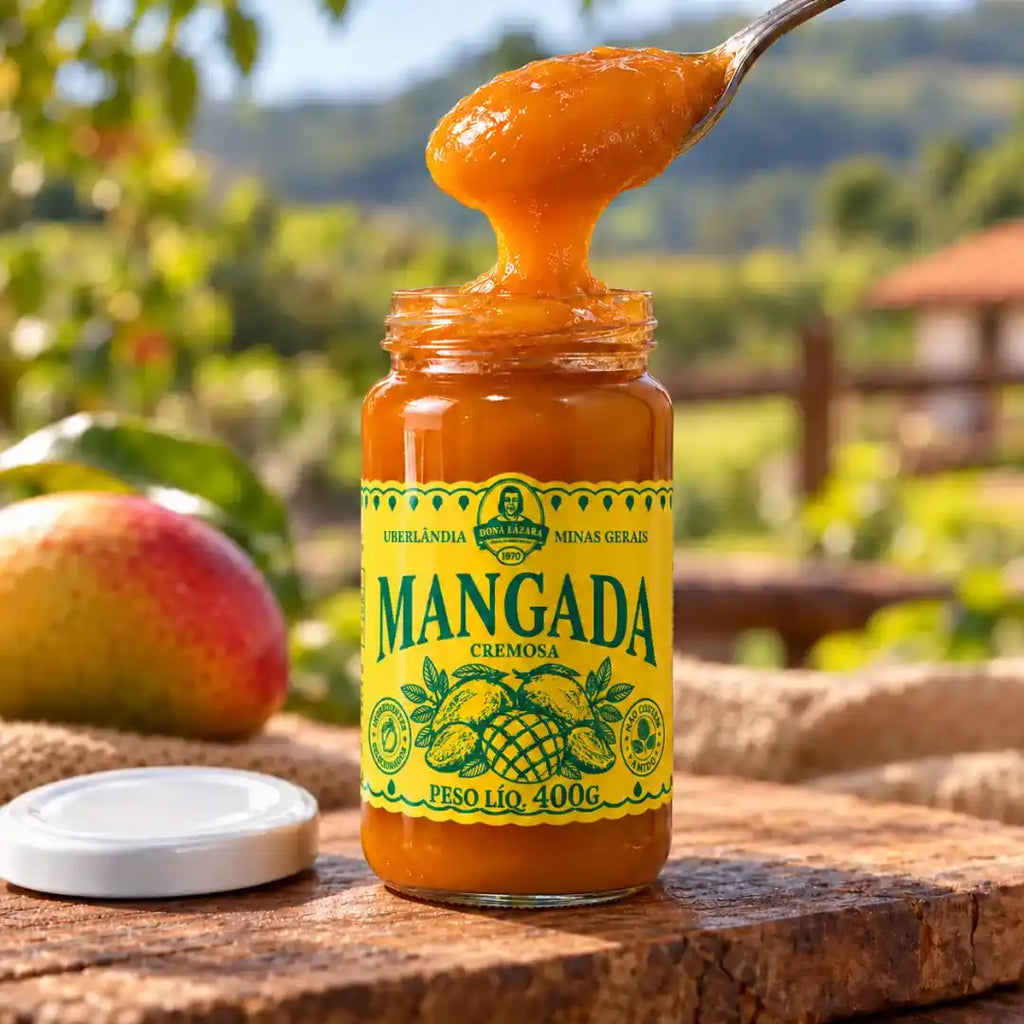 Pote aberto de mangada Dona Lázara com colher puxando o doce cremoso, tampa branca sobre a mesa e manga madura apoiada em tecido de juta.
