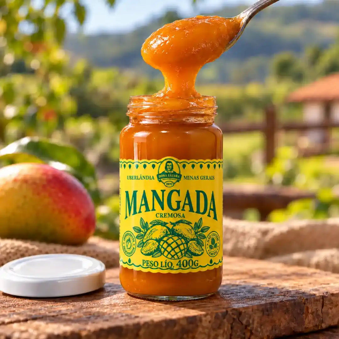 Pote aberto de mangada Dona Lázara com colher puxando o doce cremoso, tampa branca sobre a mesa e manga madura apoiada em tecido de juta.