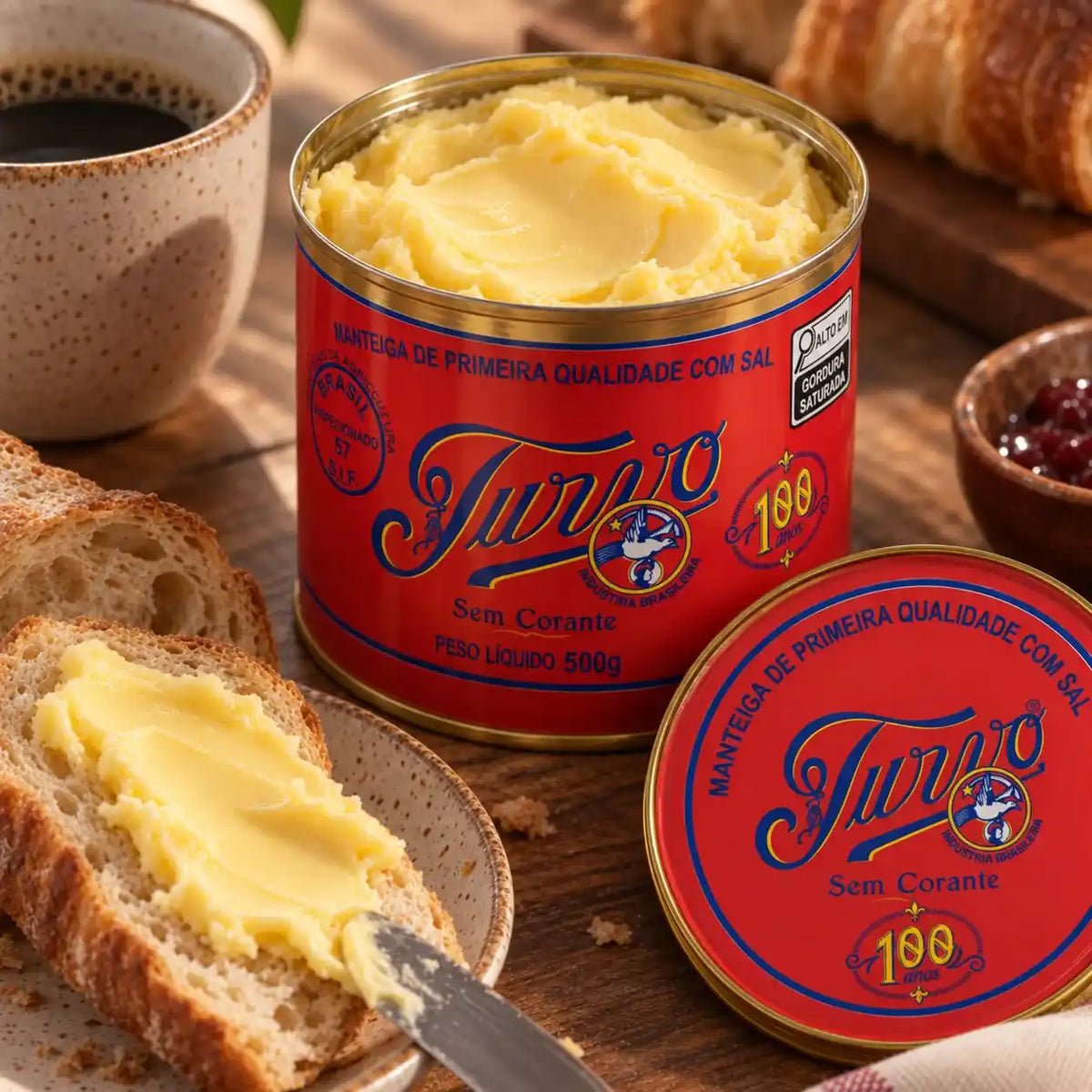 Lata aberta de Manteiga Turvo com sal com a manteiga cremosa em destaque, tampa vermelha ao lado, fatia de pão passada com manteiga, xícara de café e tigela de geleia sobre mesa rústica.