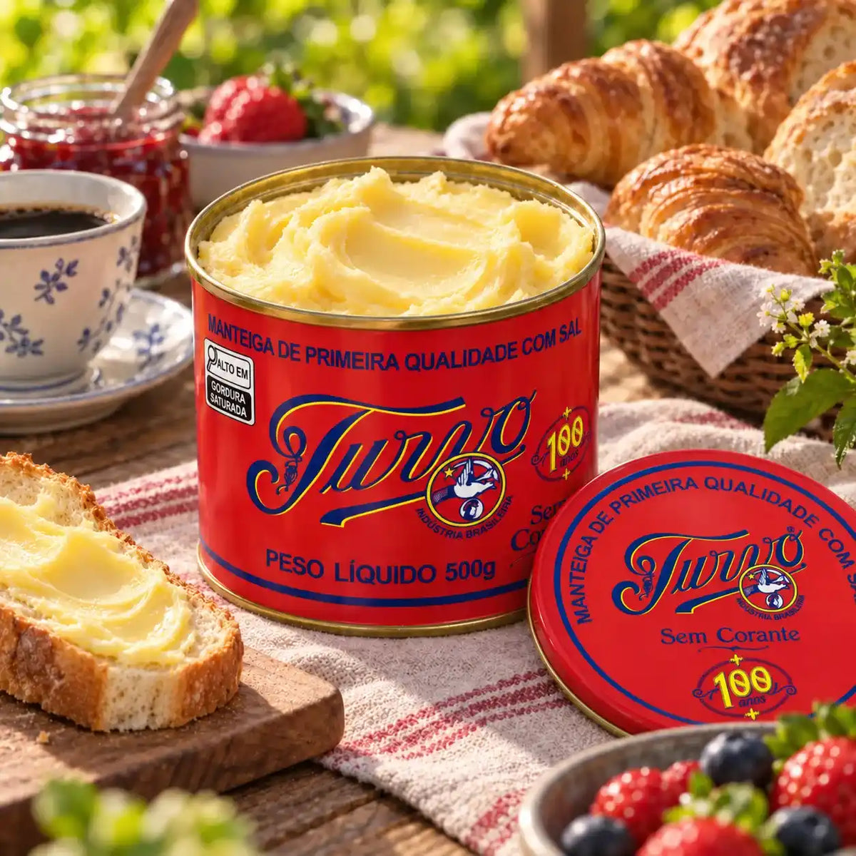 Lata aberta de Manteiga Turvo com sal com a manteiga cremosa, tampa vermelha com o selo "100 anos" apoiada ao lado, torrada passada com manteiga, croissants, geleia e frutas vermelhas numa mesa ensolarada.