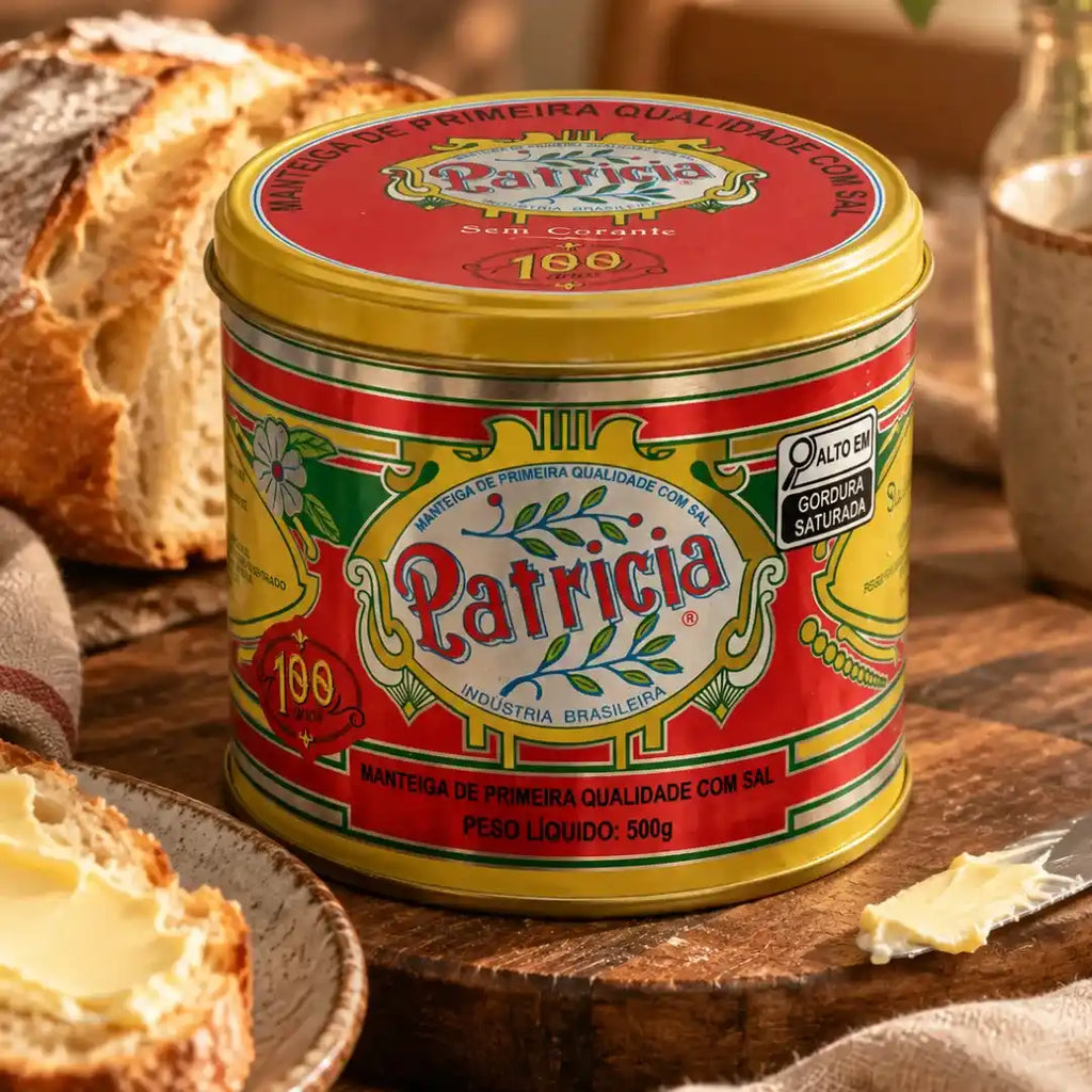 Lata fechada de Manteiga Patrícia com sal 500g mostrando a tampa e o rótulo vermelho, com o selo "alto em gordura saturada" e o peso líquido visíveis, pão artesanal e café ao redor sobre mesa rústica.