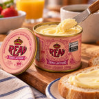 Faca tirando manteiga sem lactose da lata aberta da Real, com a tampa rosa de coroa, selo de 100 anos e os dizeres manteiga de primeira qualidade apoiada ao lado, e pão fatiado na tábua.