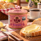 Lata fechada de Manteiga Real Zero Lactose com sal, 200g, sobre toalha xadrez vermelha, com fatia de pão passada com manteiga, faca, cesta de pães e café ao fundo.