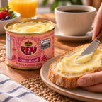 Mão passando manteiga zero lactose no pão com faca, ao lado da lata aberta da Real cheia até a borda, com xícara de café, suco de laranja e frutas vermelhas ao fundo.